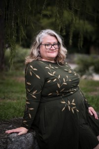 Photograph of Frances Donovan: a chubby woman in a green dress embroidered with gold leaves. She has graying, shoulder-length blonde hair and smiles at the camera. The drooping tendrils of a larch tree frame her and she sits on a stone wall.